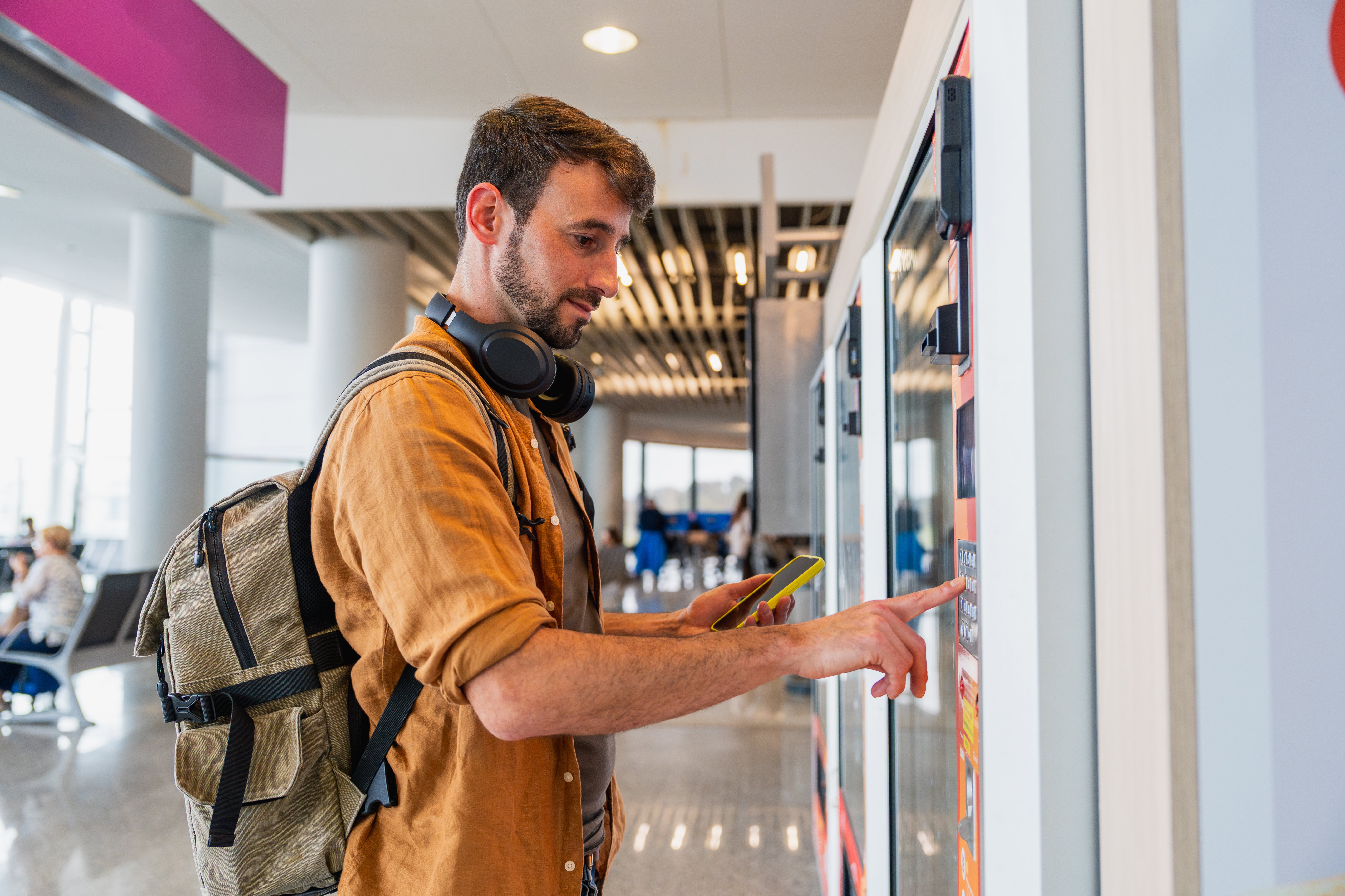 Young man using vending machine with smartphone and headphones while traveling